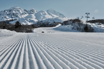 Groomed ski slopes stretch towards rugged, snow-capped mountains under a clear blue sky. Cable cars traverse overhead, positioned to the right, while trees sparsely line the lower slopes, contrasting against the pristine white snow.