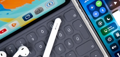 iPad with keyboard, white stylus, white AirPods; and an iphone on table