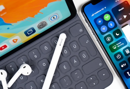 iPad with keyboard, white stylus, white AirPods; and an iphone on table