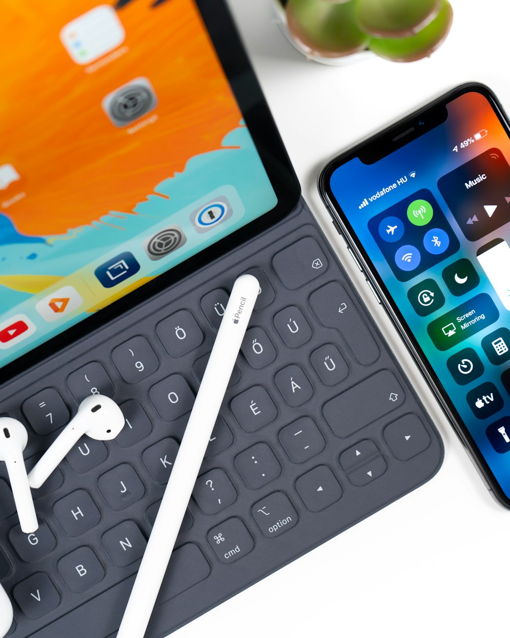 iPad with keyboard, white stylus, white AirPods; and an iphone on table