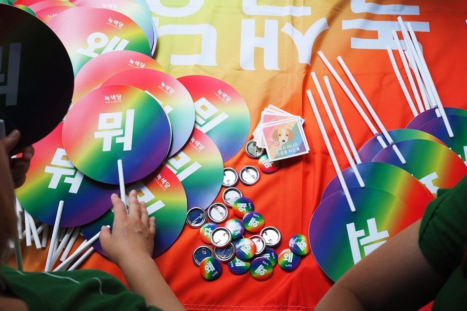 An assortment of colorful rainbow fans with Korean writing, buttons, and pamphlets arranged on a gradient orange to yellow background. People are visible interacting with the items.