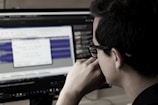 A person wearing glasses is deeply focused on a computer screen displaying audio editing software. The screen shows waveforms and various controls while the room has a dim, professional atmosphere.