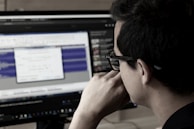 A person wearing glasses is deeply focused on a computer screen displaying audio editing software. The screen shows waveforms and various controls while the room has a dim, professional atmosphere.