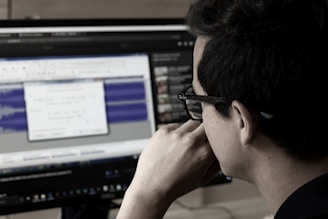A person wearing glasses is deeply focused on a computer screen displaying audio editing software. The screen shows waveforms and various controls while the room has a dim, professional atmosphere.