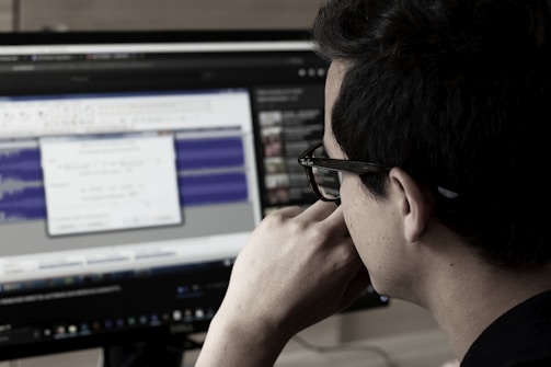 A person wearing glasses is deeply focused on a computer screen displaying audio editing software. The screen shows waveforms and various controls while the room has a dim, professional atmosphere.