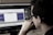 A person wearing glasses is deeply focused on a computer screen displaying audio editing software. The screen shows waveforms and various controls while the room has a dim, professional atmosphere.
