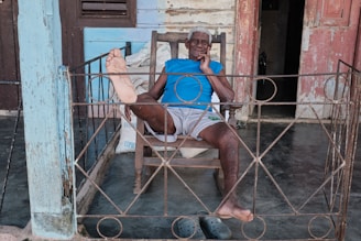 An elderly person sits in a rocking chair on a porch surrounded by an ornate metal railing. The individual is wearing a bright blue shirt and light-colored shorts, with their feet casually placed on the railing. The background features a wooden building with weathered paint in shades of blue and pink.