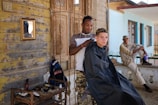 A young man is getting a haircut outdoors by a barber who is using clippers. An older man in casual clothes sits nearby on a railing, seemingly observing or resting. The setting appears rustic with weathered wooden walls, a small mirror, and various barber tools arranged on a table. The atmosphere is informal and relaxed.