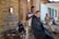 A young man is getting a haircut outdoors by a barber who is using clippers. An older man in casual clothes sits nearby on a railing, seemingly observing or resting. The setting appears rustic with weathered wooden walls, a small mirror, and various barber tools arranged on a table. The atmosphere is informal and relaxed.
