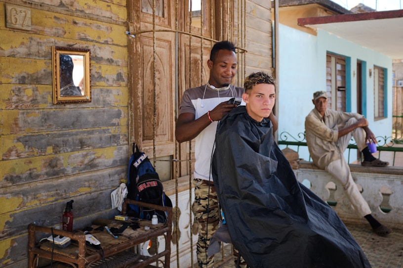 A young man is getting a haircut outdoors by a barber who is using clippers. An older man in casual clothes sits nearby on a railing, seemingly observing or resting. The setting appears rustic with weathered wooden walls, a small mirror, and various barber tools arranged on a table. The atmosphere is informal and relaxed.