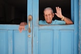 An elderly person wearing a light blue shirt is smiling and waving while leaning through a bright blue wooden door. The door has a vintage handle and a lock, and the person is wearing a wristwatch.