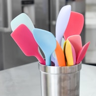 A collection of colorful silicone spatulas is arranged in a stainless steel holder. The spatulas come in a variety of pastel shades, including pink, blue, yellow, orange, and lilac. The holder is positioned on a light gray surface, likely a kitchen countertop, with blurry kitchen appliances in the background.