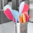 A collection of colorful silicone spatulas is arranged in a stainless steel holder. The spatulas come in a variety of pastel shades, including pink, blue, yellow, orange, and lilac. The holder is positioned on a light gray surface, likely a kitchen countertop, with blurry kitchen appliances in the background.