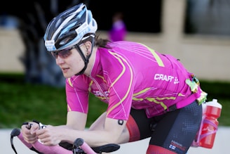 A cyclist wearing a pink jersey and black cycling shorts, equipped with a helmet and sunglasses, riding a bicycle in an aerodynamic position. A red water bottle is attached to the side of the bike, and a small item is clipped to the back of the waistband.