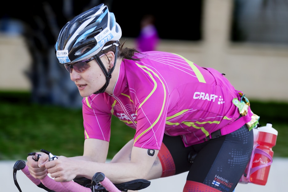 A cyclist wearing a pink jersey and black cycling shorts, equipped with a helmet and sunglasses, riding a bicycle in an aerodynamic position. A red water bottle is attached to the side of the bike, and a small item is clipped to the back of the waistband.