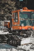 An orange tracked snow vehicle is parked outdoors on a snowy surface. The vehicle appears to be equipped for snow grooming, with a wide front windshield and a small orange light on top. The tracks are designed to help the vehicle move through snow. Trees can be seen in the background.