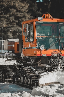 An orange tracked snow vehicle is parked outdoors on a snowy surface. The vehicle appears to be equipped for snow grooming, with a wide front windshield and a small orange light on top. The tracks are designed to help the vehicle move through snow. Trees can be seen in the background.