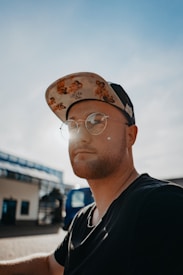 A person wearing glasses and a decorated baseball cap is captured in a close-up shot. The sun is shining brightly, creating a halo effect around their head. The background shows a clear blue sky and an out-of-focus building, giving a sense of a warm, sunny day.