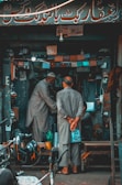 A small, cluttered shop with two men wearing traditional attire, engaged in a conversation or transaction. Various tools, equipment, and lights are visible within the shop. A motorcycle helmet is in the foreground.
