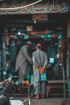A small, cluttered shop with two men wearing traditional attire, engaged in a conversation or transaction. Various tools, equipment, and lights are visible within the shop. A motorcycle helmet is in the foreground.