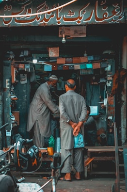 A small, cluttered shop with two men wearing traditional attire, engaged in a conversation or transaction. Various tools, equipment, and lights are visible within the shop. A motorcycle helmet is in the foreground.