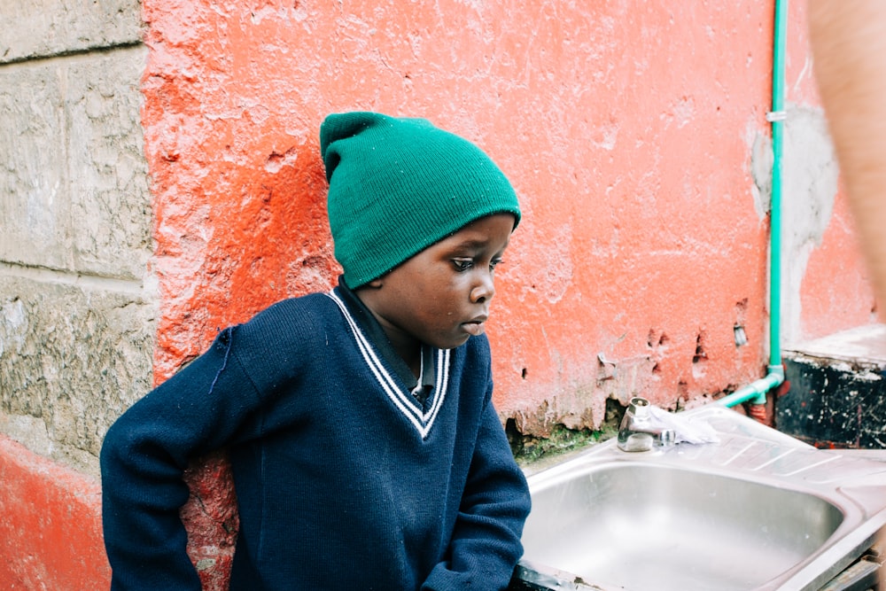 boy in green knit cap and blue sweatshirt leaning on orange painted wall beside gray stainless steel sink