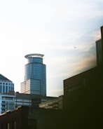 A modern urban skyline featuring a cylindrically shaped glass skyscraper standing prominently. Additional office buildings and structures are visible in the foreground. The building with the sign 'Hennepin Healthcare' appears on the right. The sky is partly cloudy, and a small airplane is seen flying in the distance.