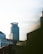 A modern urban skyline featuring a cylindrically shaped glass skyscraper standing prominently. Additional office buildings and structures are visible in the foreground. The building with the sign 'Hennepin Healthcare' appears on the right. The sky is partly cloudy, and a small airplane is seen flying in the distance.
