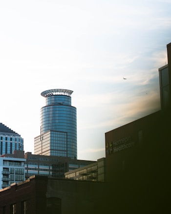 A modern urban skyline featuring a cylindrically shaped glass skyscraper standing prominently. Additional office buildings and structures are visible in the foreground. The building with the sign 'Hennepin Healthcare' appears on the right. The sky is partly cloudy, and a small airplane is seen flying in the distance.