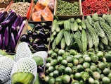 A vibrant assortment of fresh vegetables displayed neatly in a market setting. The array includes purple eggplants, small green peas, bitter gourds, chillies, green eggplants, and other fresh produce packed in netting.