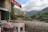 A rustic outdoor restaurant terrace with a view of mountainous landscape. The stone building has an open window with green shutters and a red awning displaying the word 'restaurant'. The seating area consists of wooden tables and chairs with red and white patterned cushions. The background features lush green hills and a cloudy sky, along with a small village nestled in the valley.