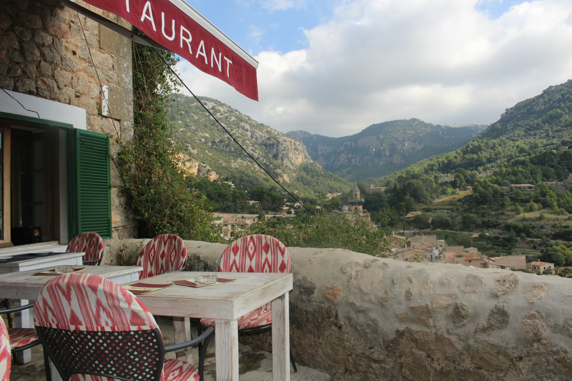 A rustic outdoor restaurant terrace with a view of mountainous landscape. The stone building has an open window with green shutters and a red awning displaying the word 'restaurant'. The seating area consists of wooden tables and chairs with red and white patterned cushions. The background features lush green hills and a cloudy sky, along with a small village nestled in the valley.