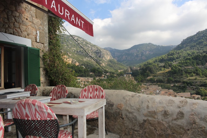 A rustic outdoor restaurant terrace with a view of mountainous landscape. The stone building has an open window with green shutters and a red awning displaying the word 'restaurant'. The seating area consists of wooden tables and chairs with red and white patterned cushions. The background features lush green hills and a cloudy sky, along with a small village nestled in the valley.