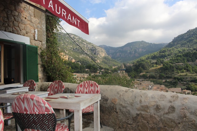 A rustic outdoor restaurant terrace with a view of mountainous landscape. The stone building has an open window with green shutters and a red awning displaying the word 'restaurant'. The seating area consists of wooden tables and chairs with red and white patterned cushions. The background features lush green hills and a cloudy sky, along with a small village nestled in the valley.