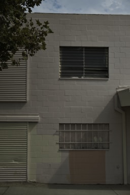 A plain, rectangular brick wall painted in a muted beige color. There is a single window with horizontal blinds and a frosted lower portion. The wall is intersected by a corrugated metal door on the left and partial shadows cast from a tree. A pale pinkish patch is visible near the ground level.
