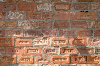 A brick wall with various engraved markings, including letters and numbers. The bricks vary in color, displaying different shades of orange and red with some weathered textures.