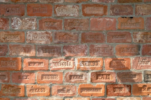 A brick wall with various engraved markings, including letters and numbers. The bricks vary in color, displaying different shades of orange and red with some weathered textures.