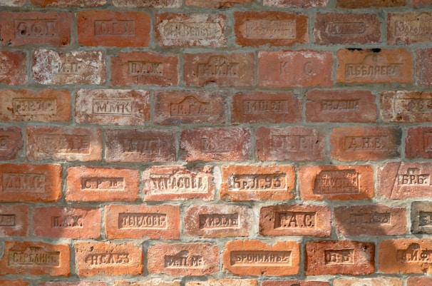 A brick wall with various engraved markings, including letters and numbers. The bricks vary in color, displaying different shades of orange and red with some weathered textures.