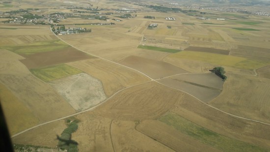 An aerial view of a patchwork landscape consisting of various agricultural fields with different shades of brown, green, and yellow. The fields are divided by dirt paths, and a few clusters of buildings are visible in the distance near the top of the image. A lone tree stands near the bottom corner.
