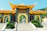 A large, ornate entrance gate with traditional East Asian architectural elements, featuring a yellow tiled roof and decorative engravings. Flanked by statues, the gateway opens to a path lined with lush greenery and leads to a set of stairs. The background includes a hillside covered in trees under a clear sky.