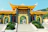 A large, ornate entrance gate with traditional East Asian architectural elements, featuring a yellow tiled roof and decorative engravings. Flanked by statues, the gateway opens to a path lined with lush greenery and leads to a set of stairs. The background includes a hillside covered in trees under a clear sky.