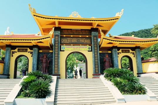 A large, ornate entrance gate with traditional East Asian architectural elements, featuring a yellow tiled roof and decorative engravings. Flanked by statues, the gateway opens to a path lined with lush greenery and leads to a set of stairs. The background includes a hillside covered in trees under a clear sky.