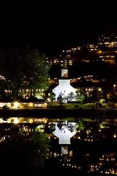 A white church is illuminated at night, situated by a body of water that reflects its image. Trees and houses are visible in the background, with small lights from the buildings creating a warm glow. The scene conveys a calm, serene night view, emphasizing the church and its mirrored reflection in the water.