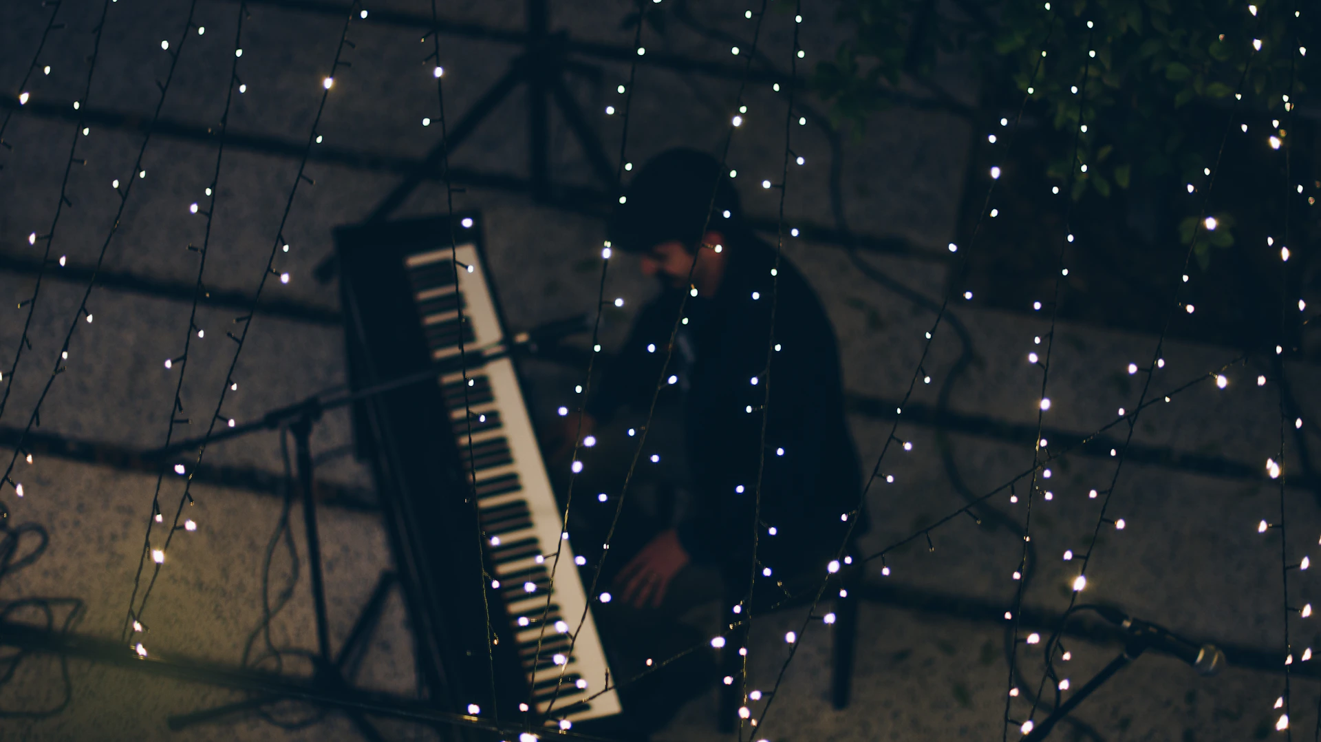 A serene outdoor setting at dusk with a pianist performing under string lights, surrounded by a small, attentive crowd seated on blankets.