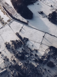 An aerial view of a snow-covered rural landscape with scattered farmhouses and buildings. Fields are neatly divided and blanketed in snow, with a network of roads connecting the properties. A small forested area provides contrast to the open fields.