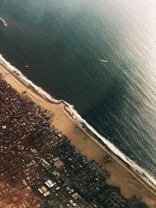 An aerial view of a coastal city with urban development on the left and a sandy beach meeting the ocean on the right. The ocean is vast, with waves gently touching the shoreline. The city appears densely populated, with numerous buildings and streets visible.