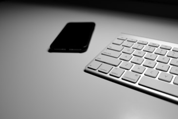 A minimalistic setup featuring a sleek keyboard on the right side with part of a smartphone visible in the upper left. The lighting adds a sense of simplicity and elegance to the scene.