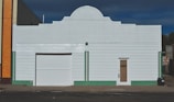 A white, rectangular building with a central roof arch, featuring a large garage door and a smaller door labeled 'Binder.' Vertical green tile lines accent the structure, contrasting with the white horizontal siding. An adjacent building with orange tiles is partially visible on the left.