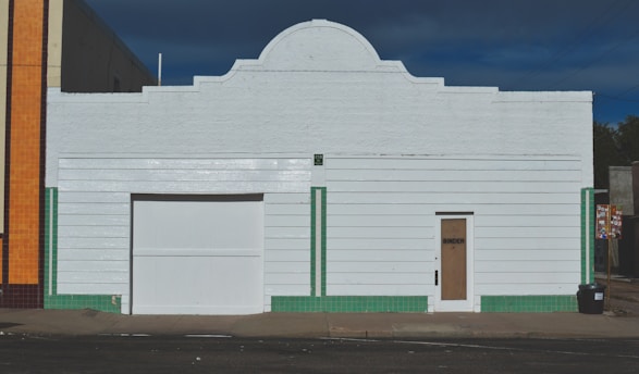 A white, rectangular building with a central roof arch, featuring a large garage door and a smaller door labeled 'Binder.' Vertical green tile lines accent the structure, contrasting with the white horizontal siding. An adjacent building with orange tiles is partially visible on the left.