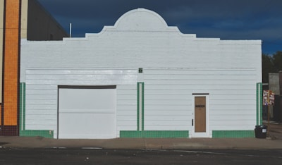 A white, rectangular building with a central roof arch, featuring a large garage door and a smaller door labeled 'Binder.' Vertical green tile lines accent the structure, contrasting with the white horizontal siding. An adjacent building with orange tiles is partially visible on the left.